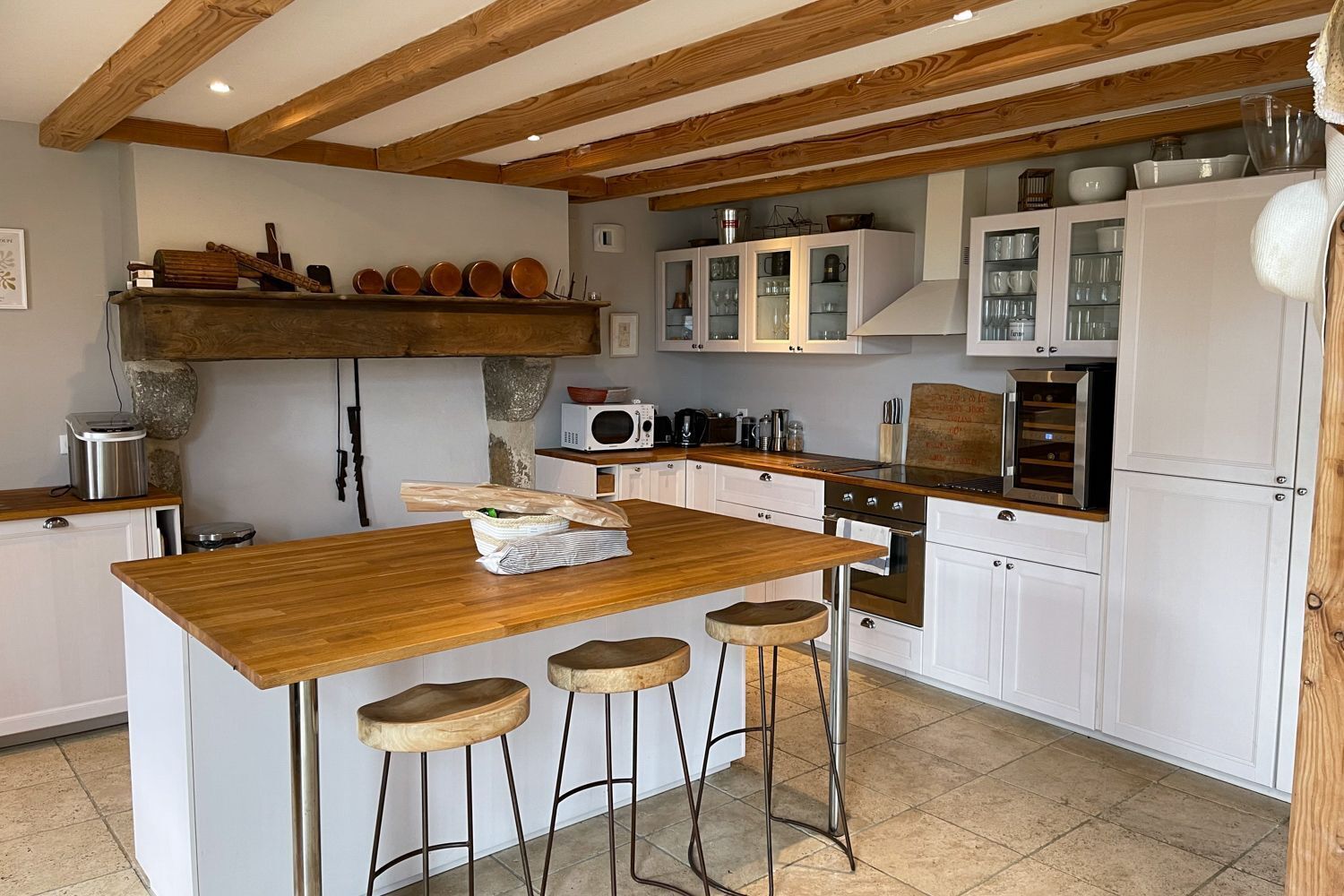 A kitchen with wooden beams, white cabinets, a central island with stools, and various kitchen appliances.