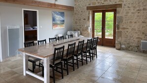 A spacious dining area with a long wooden table and chairs, a stone wall, and a large window with a view of greenery.