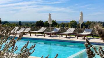 A swimming pool with lounge chairs and umbrellas, surrounded by greenery and a clear sky.