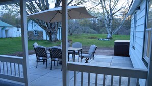 A covered patio with a table and chairs, a large umbrella, and a view of a yard with a tree and a house in the background.