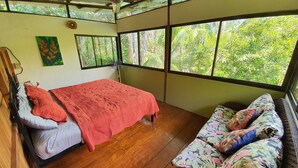 A room with a bed, a sofa, and large windows offering a view of greenery.