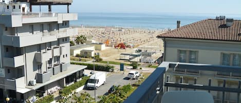 A balcony with a white chair, overlooking a beach with buildings and a clear sky.