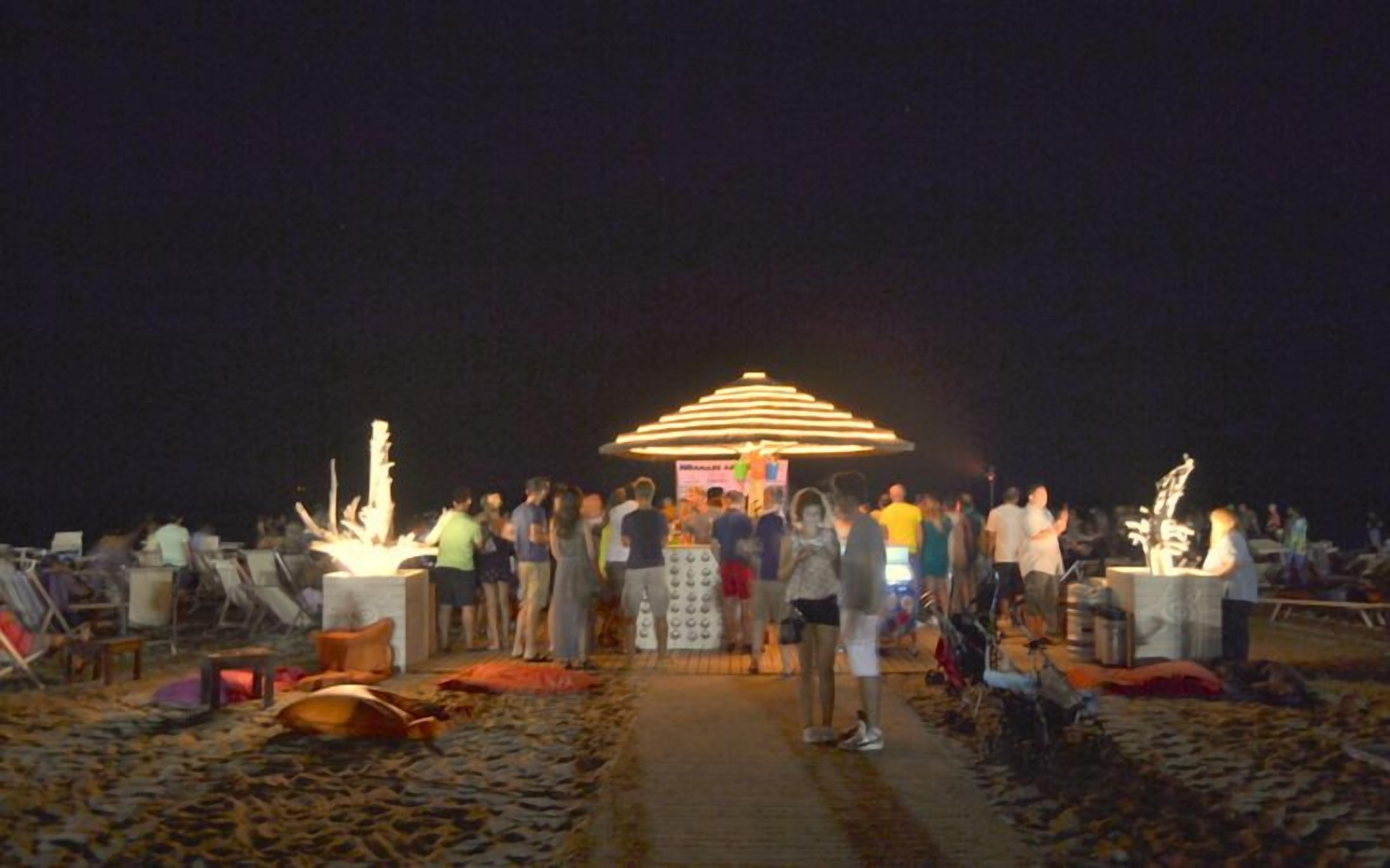 A beachside gathering with a central illuminated structure, people standing around, and various items on the ground.