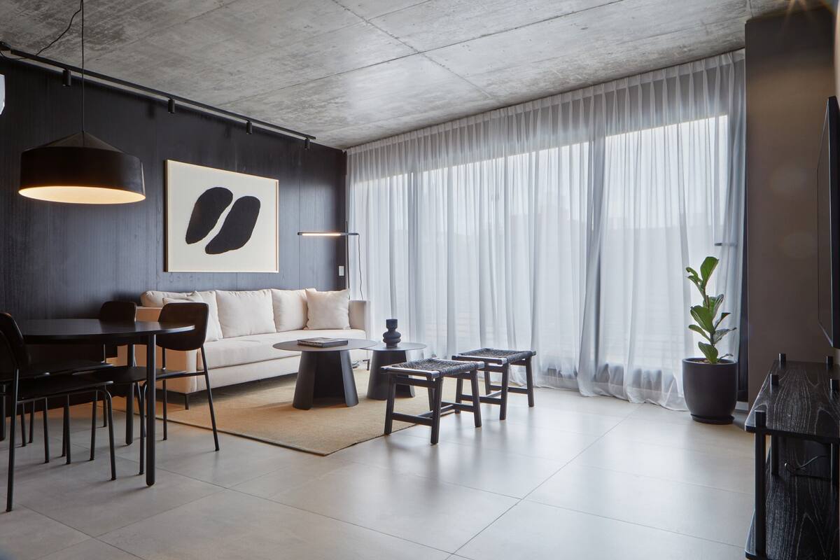 A modern living room with a sofa, dining table, chairs, and a coffee table. There is a large window with sheer curtains, a potted plant, and a framed artwork on the wall.