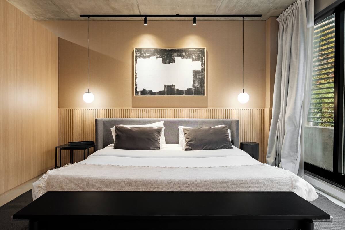 A modern bedroom with a large bed, two bedside tables, a black bench, a cityscape artwork on the wall, and a window with curtains.