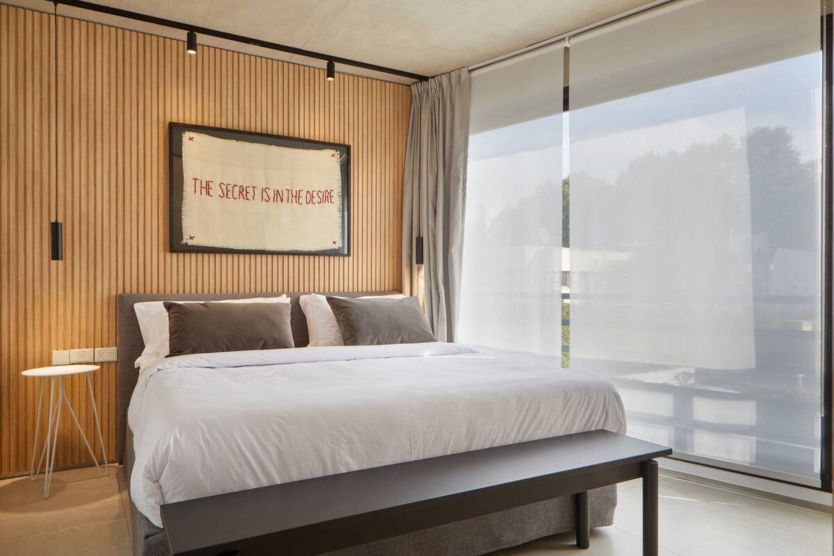 A modern bedroom with a large bed, a bench, a small table, and a wall-mounted sign.