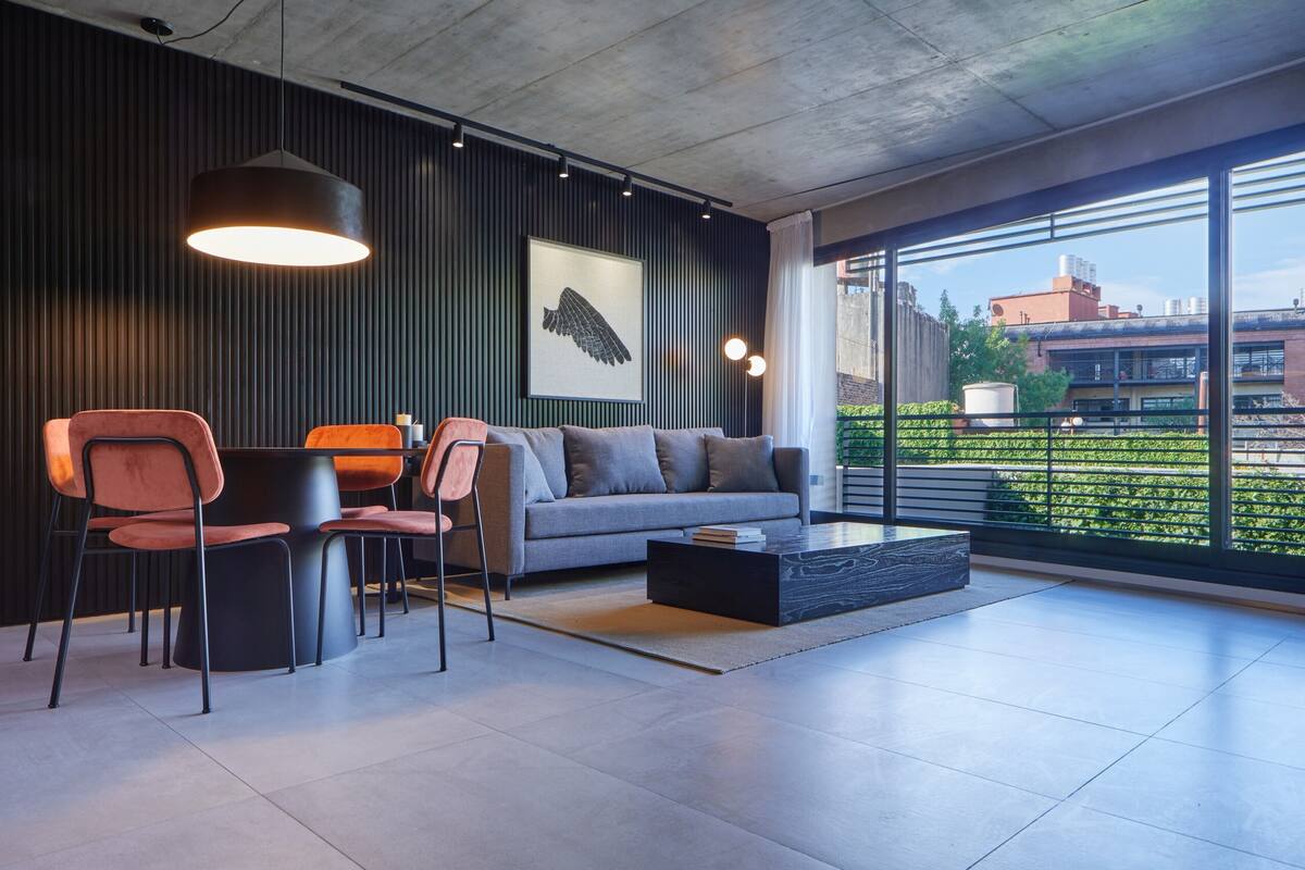 A modern living room with a sofa, chairs, and a coffee table. There is a large window with a view of buildings and greenery outside.