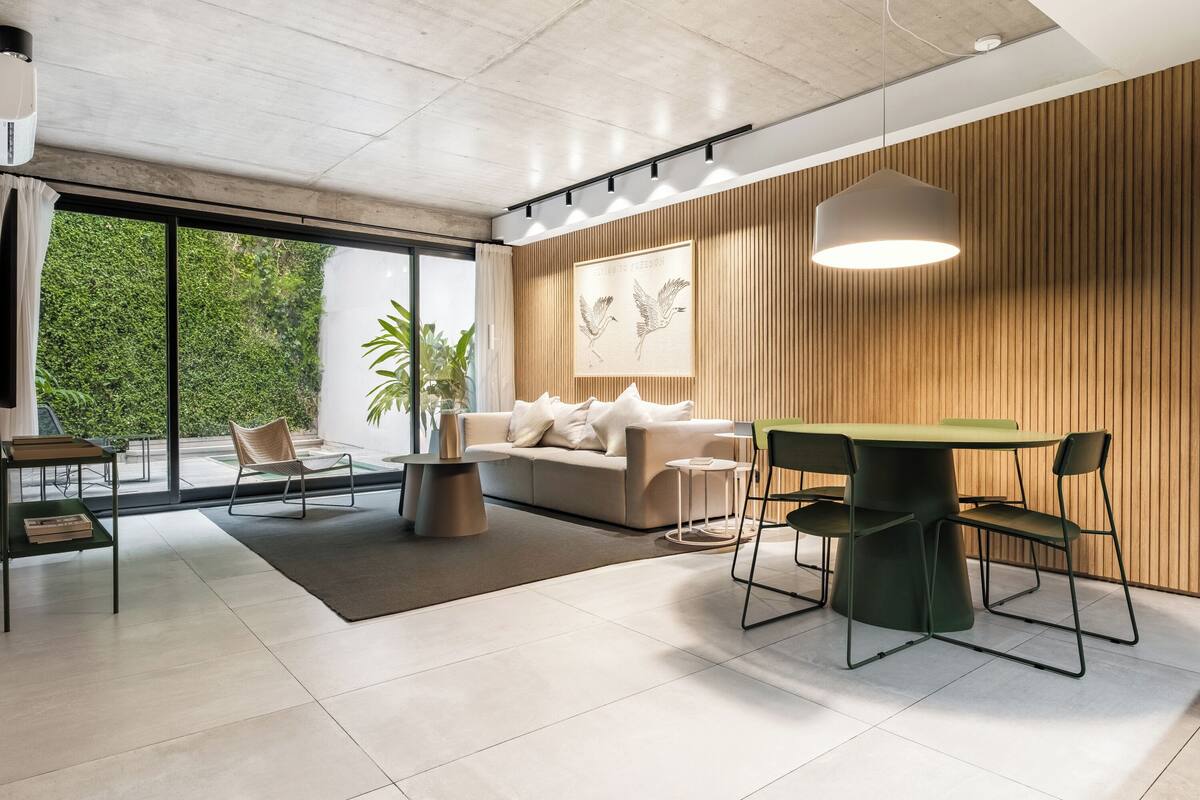 A modern living room with a sofa, dining table, chairs, and a large window offering a view of greenery.