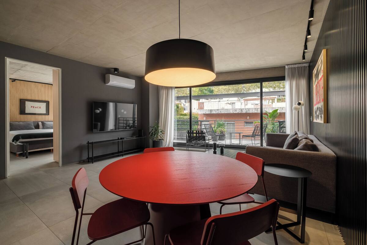 A modern living room with a round red table, black chairs, a large window with a view, a grey sofa, and a black pendant light.