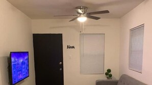 Un salon comprenant une télévision, un ventilateur de plafond, un canapé, une table à manger avec des assiettes, et une porte sur laquelle est inscrit « Clés ».