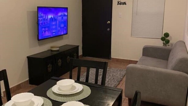 Un coin repas avec une table dressée pour quatre personnes, un téléviseur fixé au mur, un ventilateur de plafond et une porte portant l’inscription « Cuisine ».