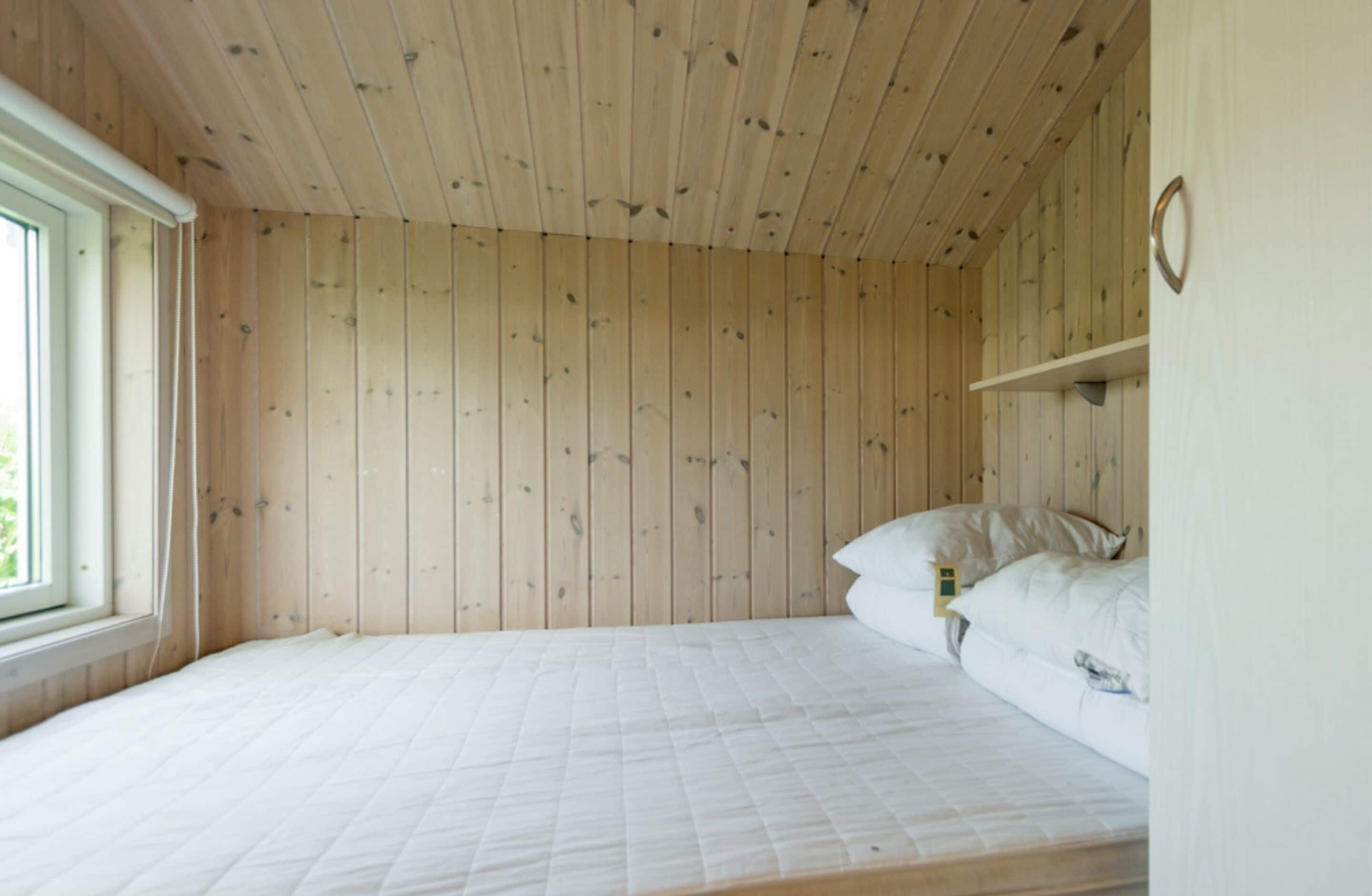 Une pièce avec un plafond et des murs recouverts de lambris en bois, un lit simple avec du linge de lit blanc et une petite étagère sur laquelle se trouve une horloge.