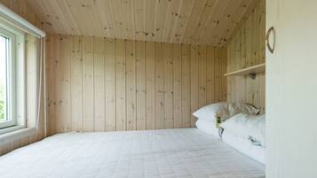 Une pièce avec un plafond et des murs recouverts de lambris en bois, un lit simple avec du linge de lit blanc et une petite étagère sur laquelle se trouve une horloge.