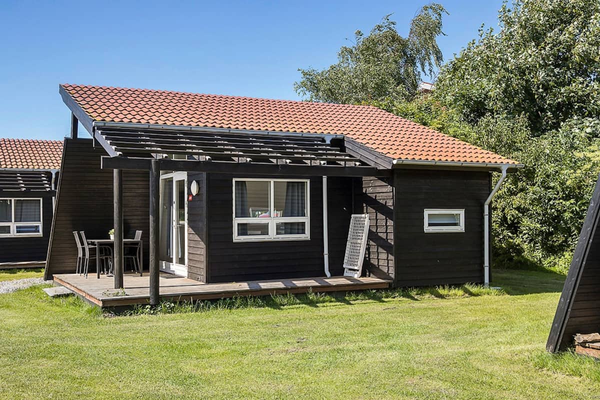 A small, dark-colored cabin with a tiled roof, a covered patio with chairs, and a window.