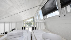 Una habitación con dos camas, ropa de cama blanca, techo inclinado y ventanas amplias con persianas.