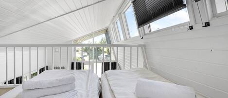 Una habitación con dos camas, ropa de cama blanca, techo inclinado y ventanas amplias con persianas.