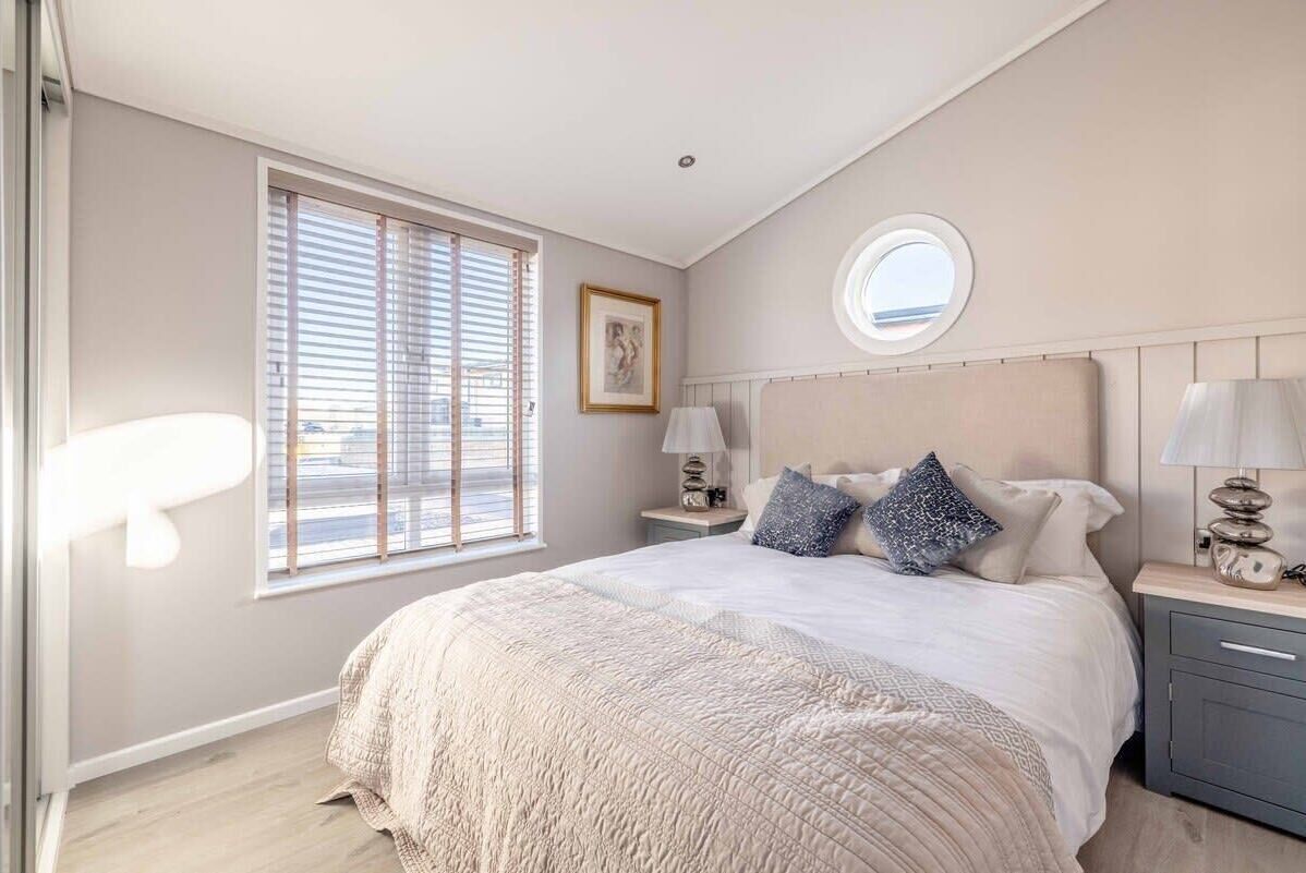 Un dormitorio con cama, mesitas de noche, ventana con persianas, tragaluz circular y un cuadro en la pared.