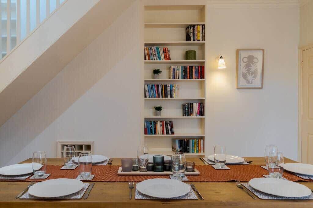 A dining area with a long table set for four, surrounded by bookshelves, a framed picture, and a staircase.