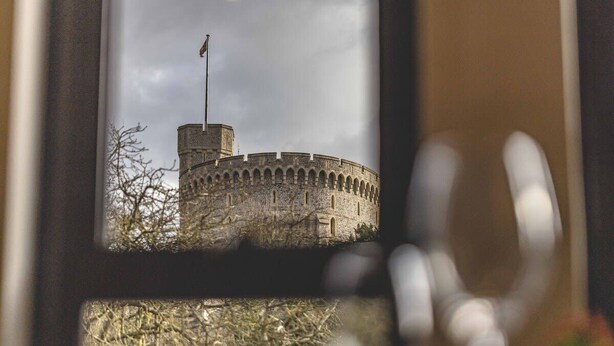 A view of a historic stone castle through a window, with a flag on top and bare trees in the foreground. - Windsor Castle Views & Parking - By Tempstay (Berkshire)