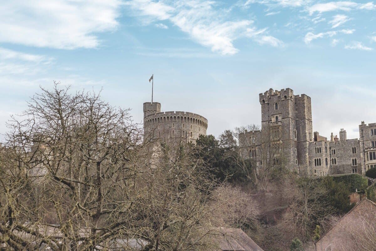 A historic castle with a round tower and a tall square tower, surrounded by trees and buildings.