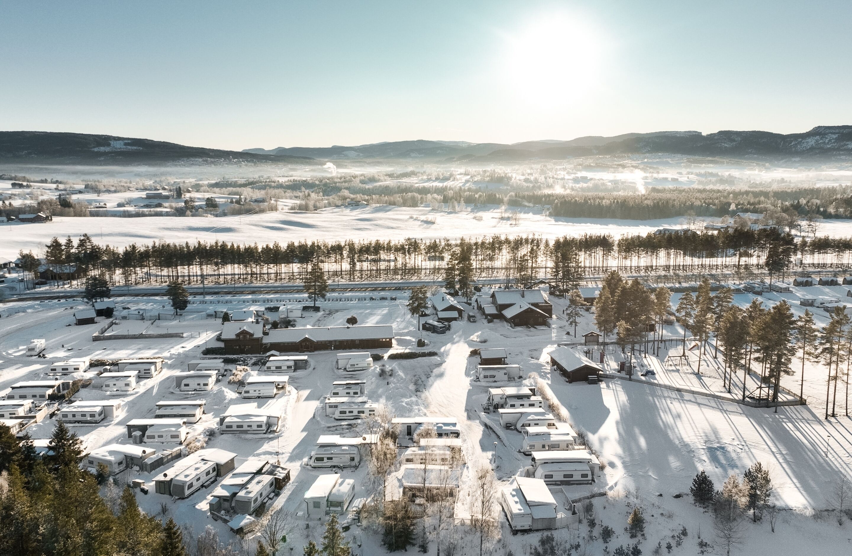 Un parque para vehículos recreativos cubierto de nieve, con varias caravanas estacionadas en filas, rodeado de árboles y colinas al fondo.