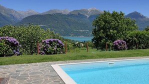Una piscina con borde de piedra, rodeada de exuberante follaje y flores moradas, con montañas al fondo.