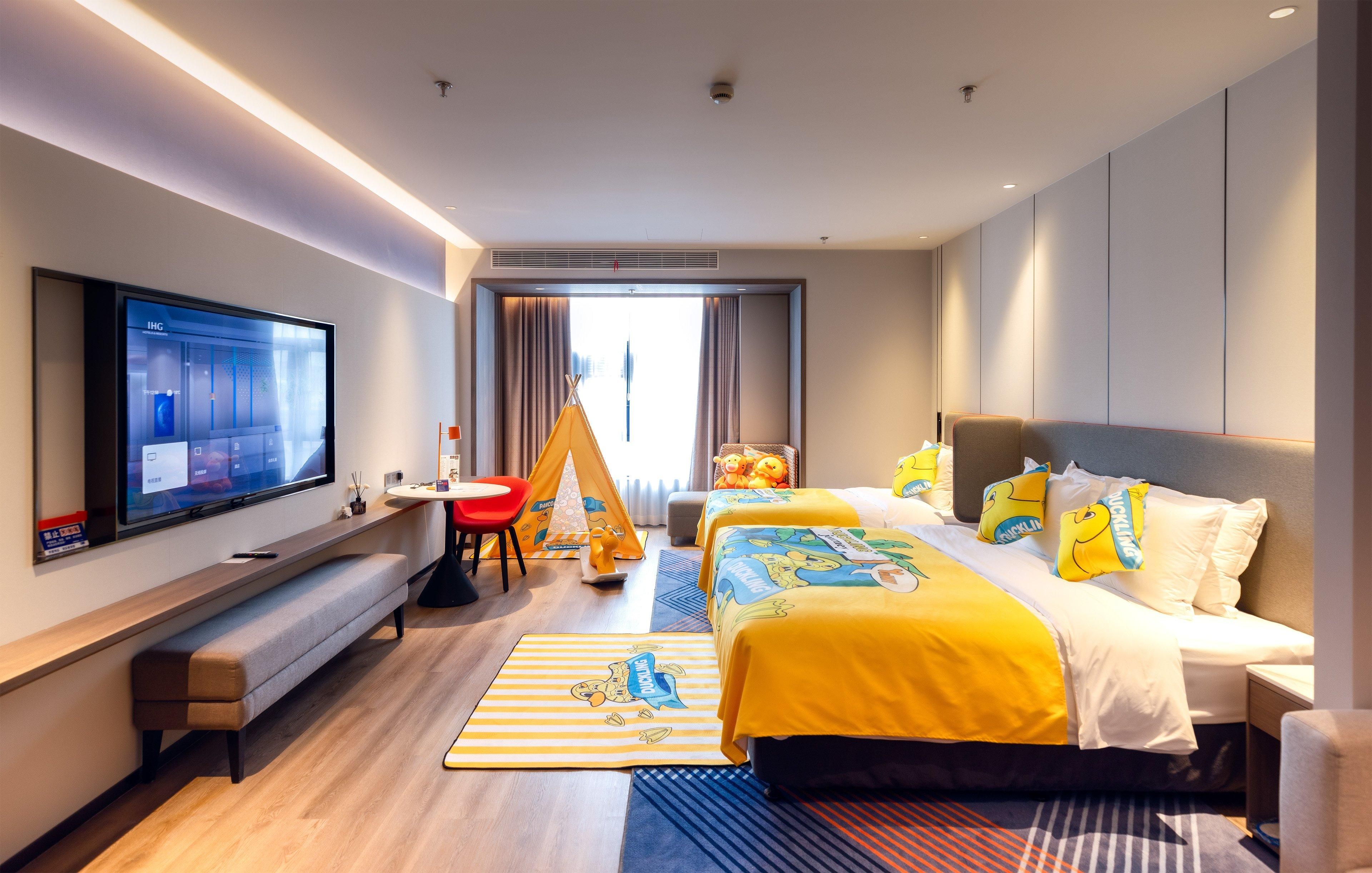 Quarto de hotel com uma TV de tela plana grande, um banco, uma cama com roupa de cama amarela, uma cadeira vermelha e uma tenda.