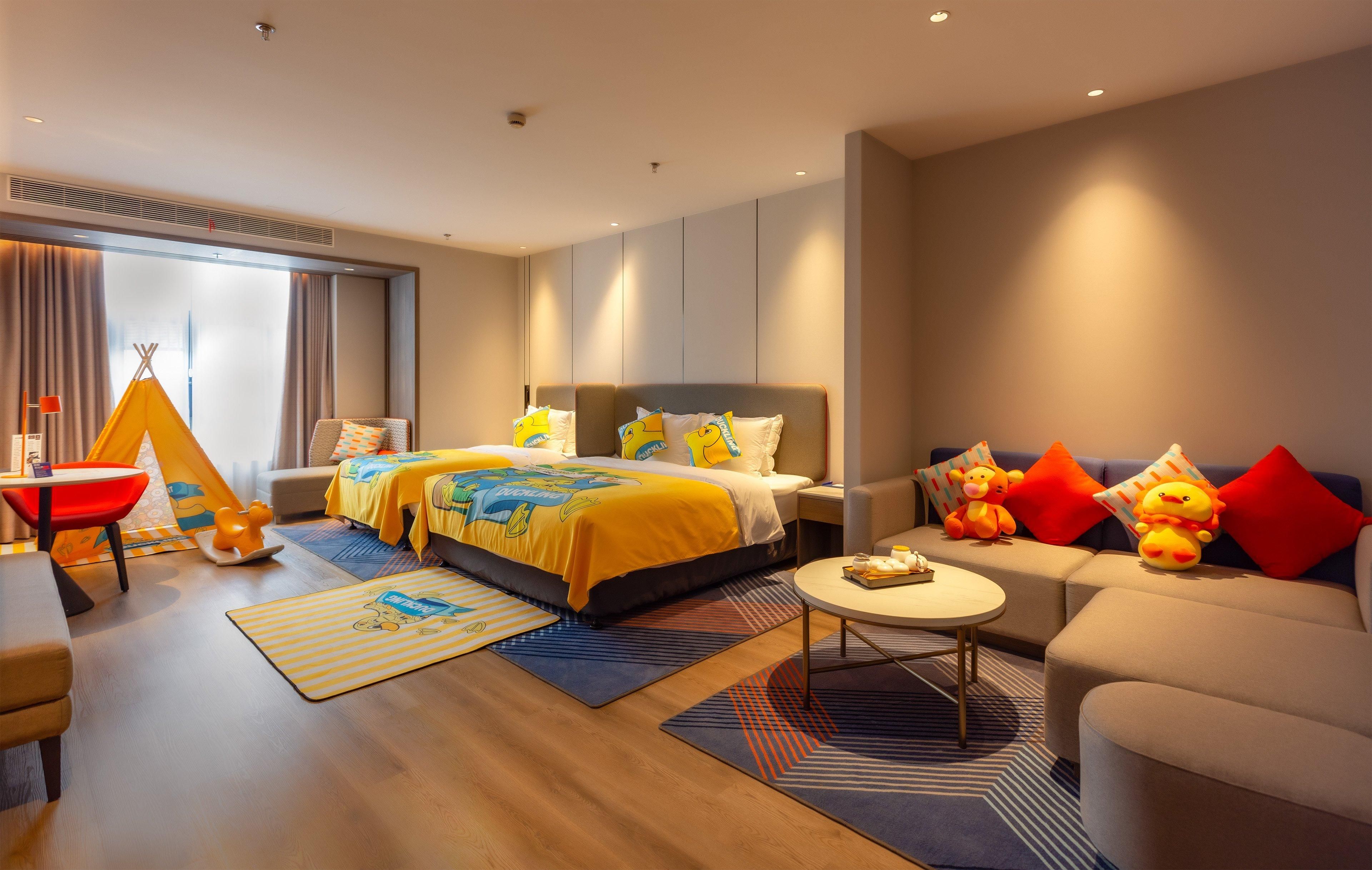 Quarto de hotel moderno com uma cama grande, um sofá, uma mesinha e um espaço de brincadeira com um tipi e brinquedos macios.