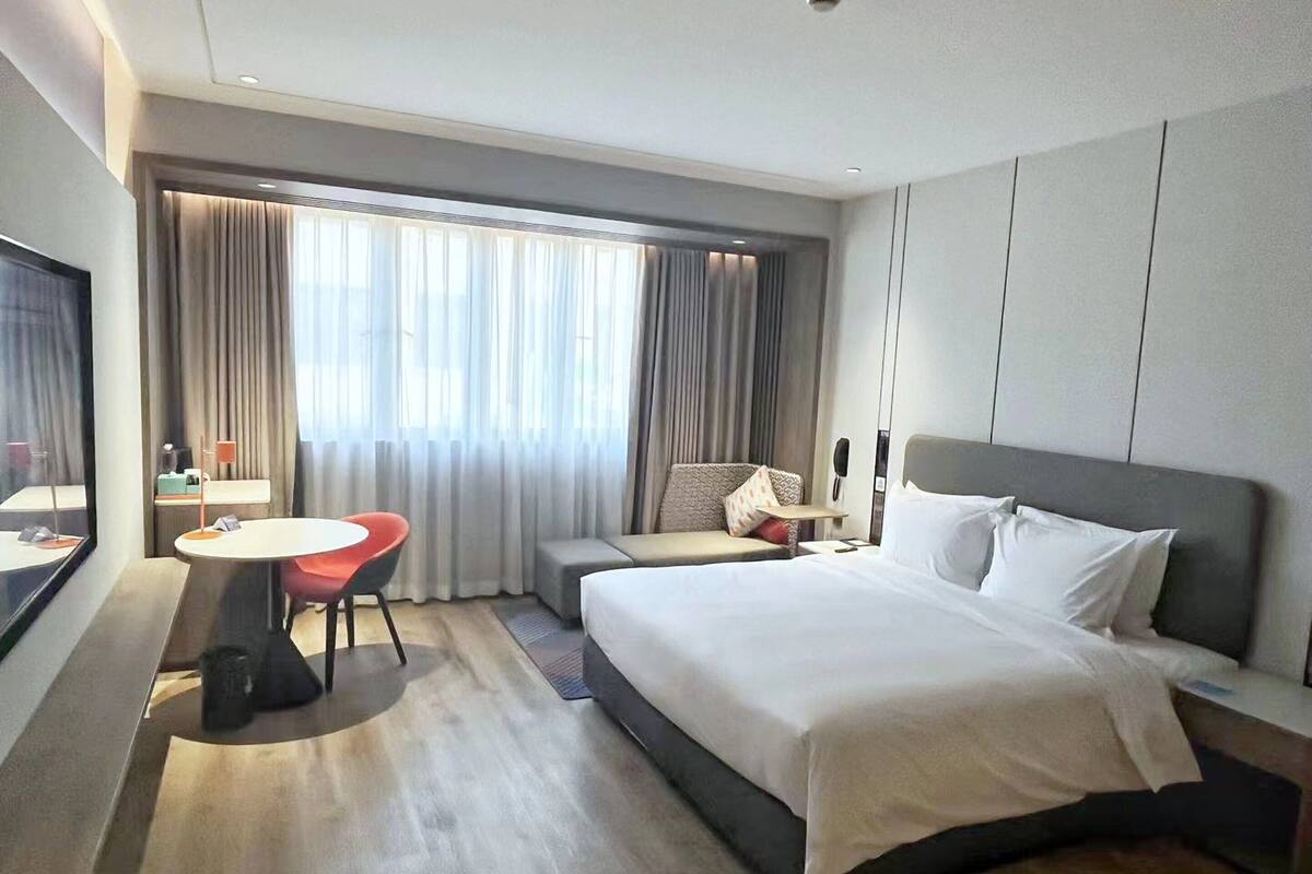 Quarto de hotel com cama, escrivaninha com cadeira, mesa pequena e janela com cortinas.