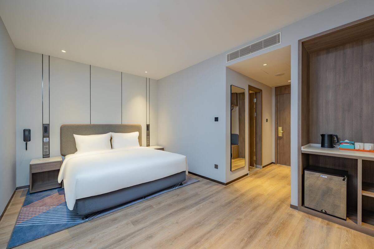 Quarto de hotel moderno com cama, mesa de cabeceira e um pequeno frigorífico.