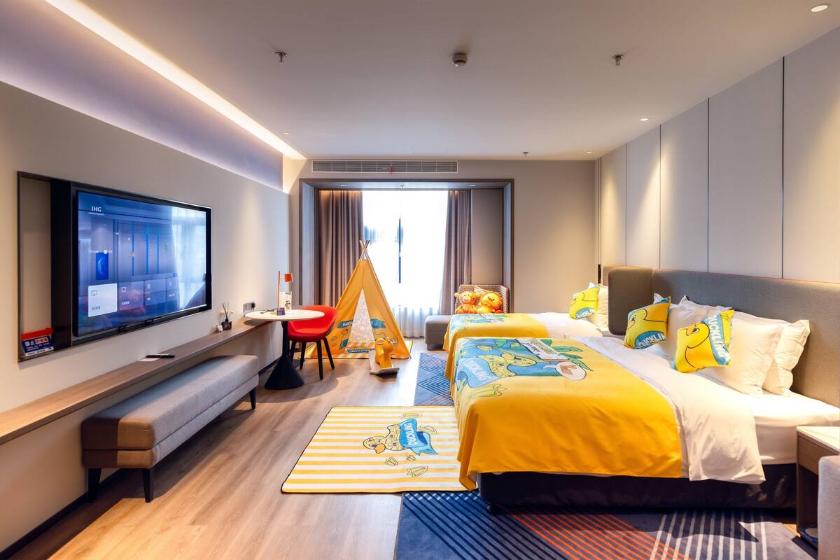 Quarto de hotel com uma TV de tela plana grande, um banco, uma cama com roupa de cama amarela, uma cadeira vermelha e uma tenda.