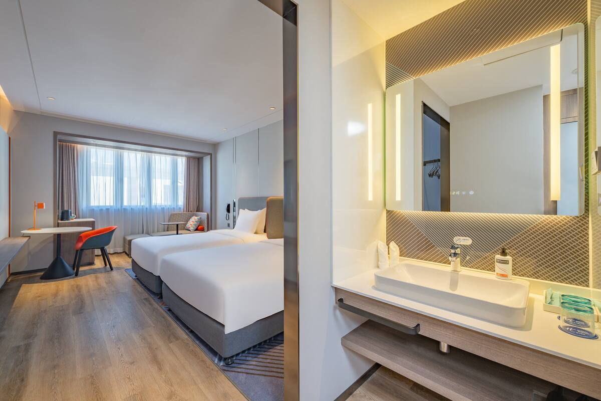 Quarto de hotel com cama, escrivaninha, cadeira e um espelho grande sobre a pia.