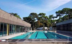 Una piscina moderna de aguas azul cristalinas, rodeada de árboles y un edificio de madera con grandes ventanales.