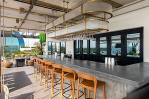 A modern bar area with a long counter, wooden stools, and a large hanging light fixture. - Dolce BY Wyndham Hollywood (HOLLYWOOD)
