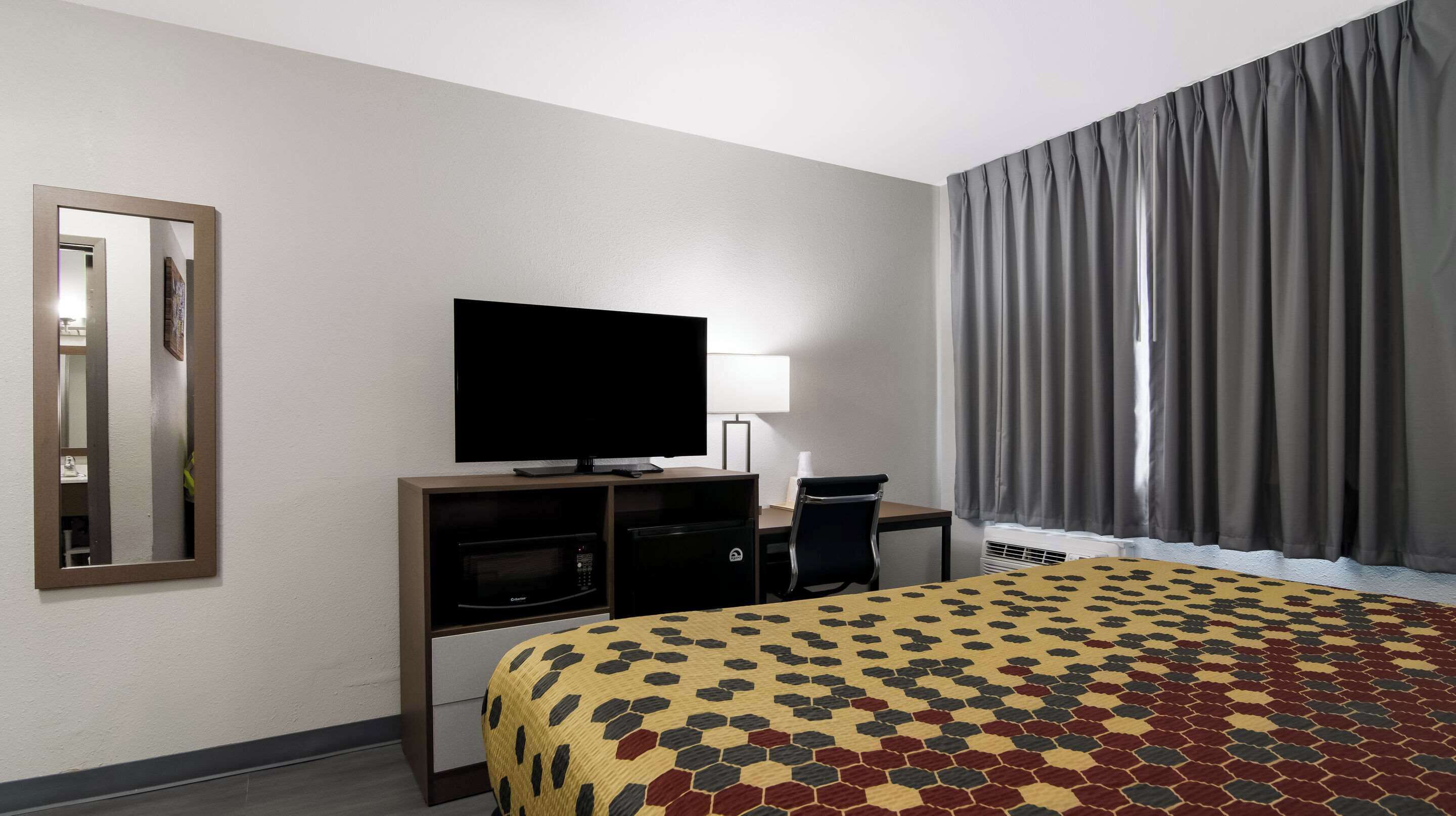 A hotel room with a bed, a television, a desk with a chair, a mirror, and a window with curtains.