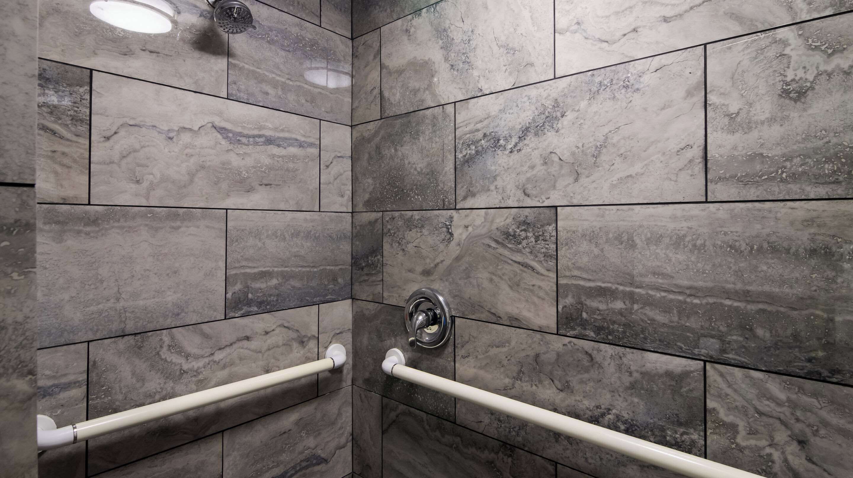 Zona de ducha con azulejos de mármol gris, un cabezal de ducha manual y dos barras de apoyo blancas.