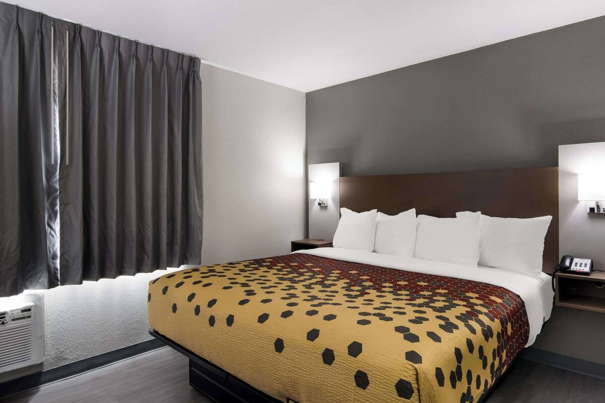 A hotel room with a large bed, grey curtains, a headboard with wall-mounted lights, a bedside table with a phone, and a wall-mounted air conditioner.