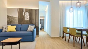 A modern living room with a blue sofa, a wooden coffee table, and yellow chairs. There is a dining area with a wooden table and chairs. A large wall art depicting an architectural scene is visible. - Tandem Populo (Cádiz)