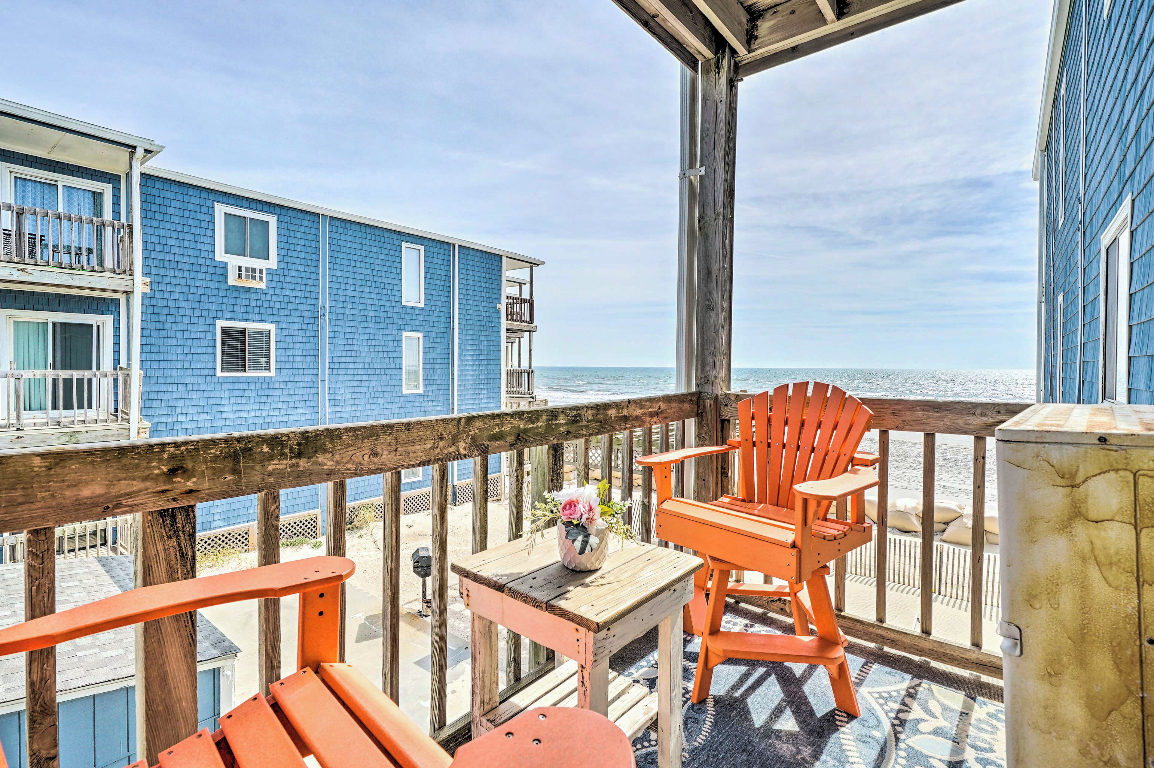 A wooden deck with orange chairs and a table, overlooking a beach with a blue building in the background.