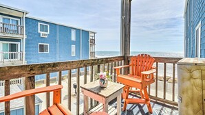 A wooden deck with orange chairs and a table, overlooking a beach with a blue building in the background. - North Topsail Condo - Steps to Beach! (North Topsail Beach)