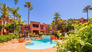 Área de piscina de resort com piscina azul cristalina, palmeiras e um edifício vermelho com varandas.