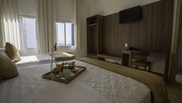 Quarto de hotel com uma cama grande, mesa de cabeceira com bandeja de café da manhã, guarda-roupa e televisão fixada na parede.