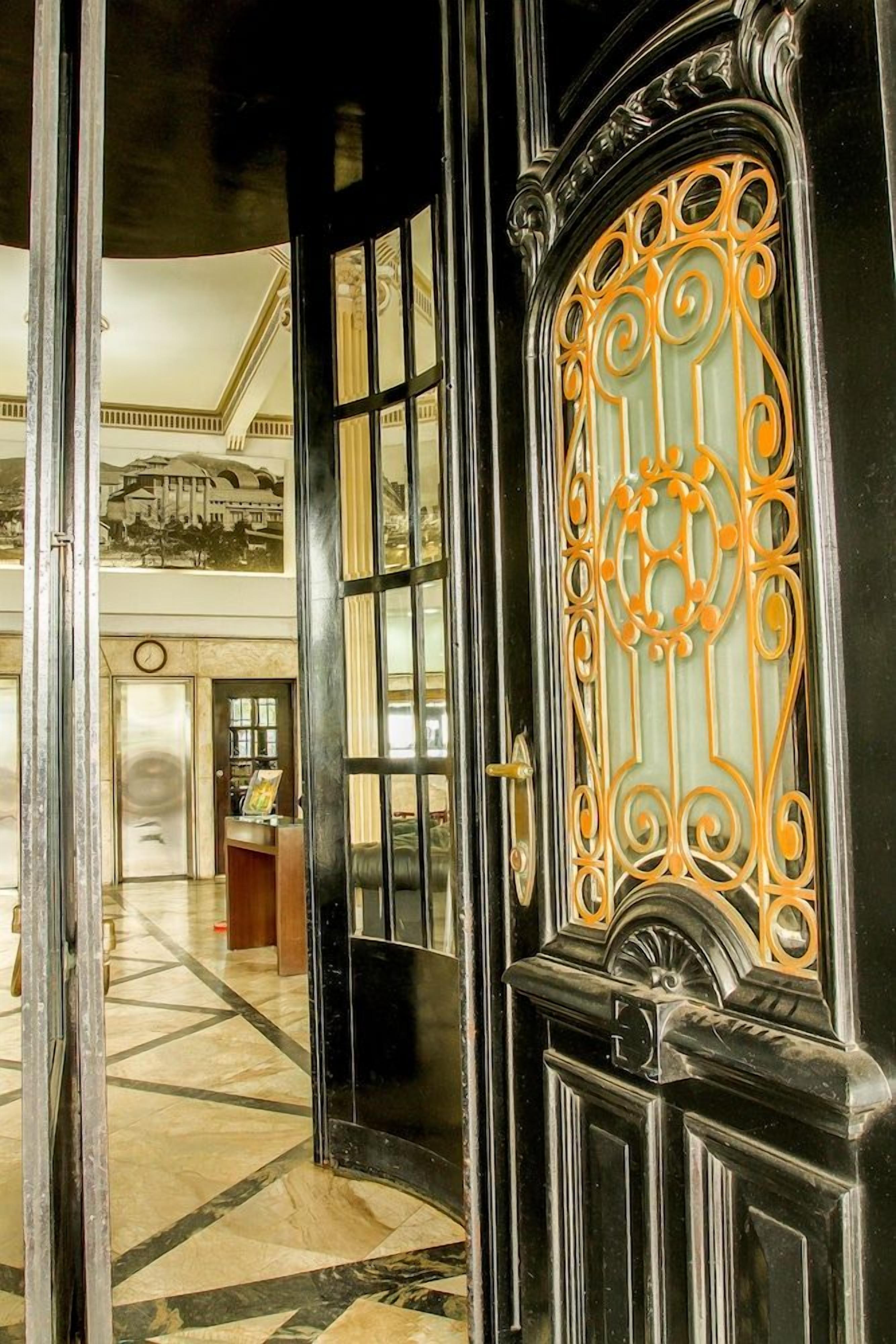 Uma porta preta ornamentada com desenhos dourados complexos, que dá acesso a um saguão com piso xadrez e um relógio na parede.