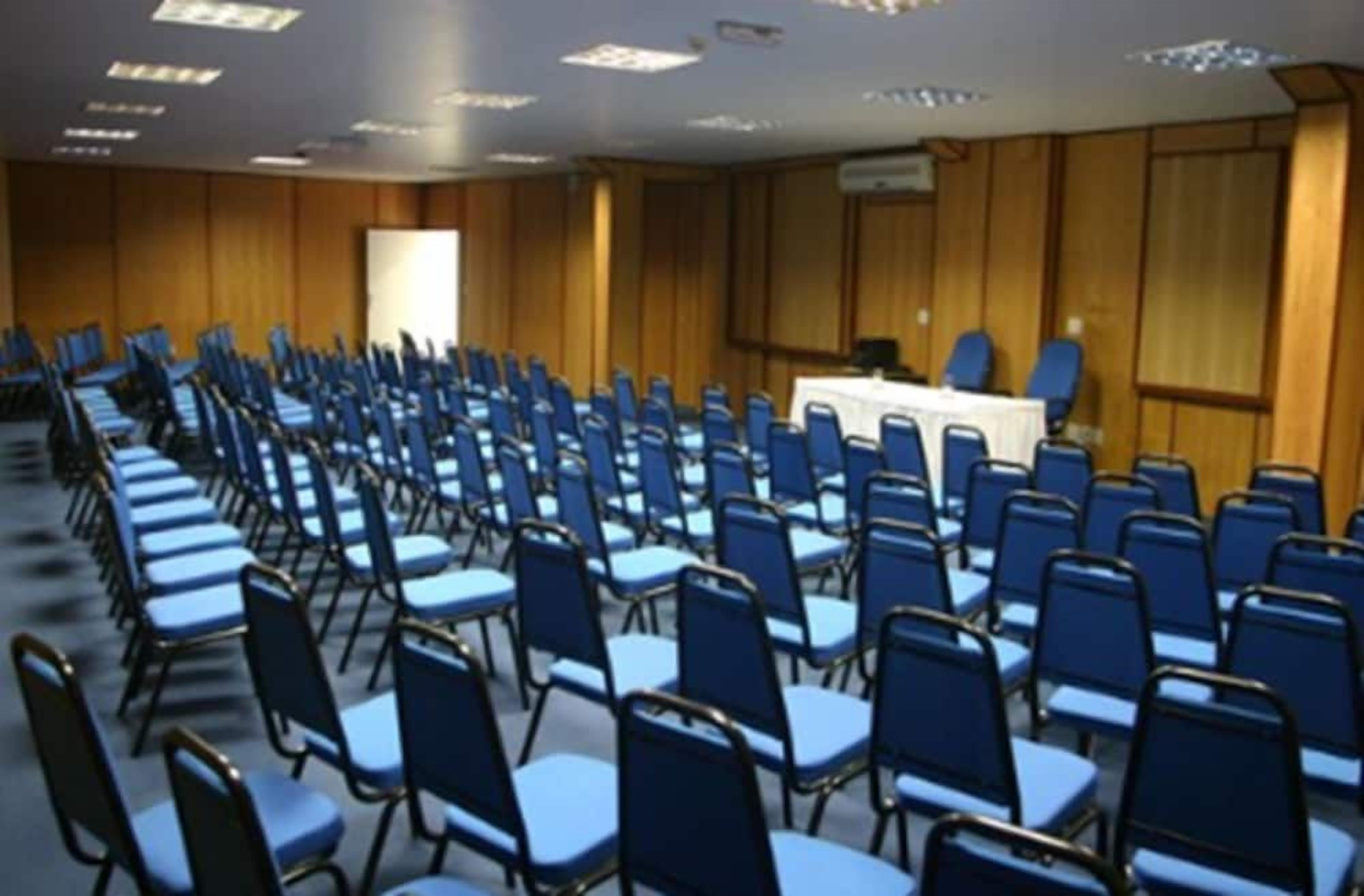 Uma sala de conferências com fileiras de cadeiras azuis e uma mesa branca à frente.
