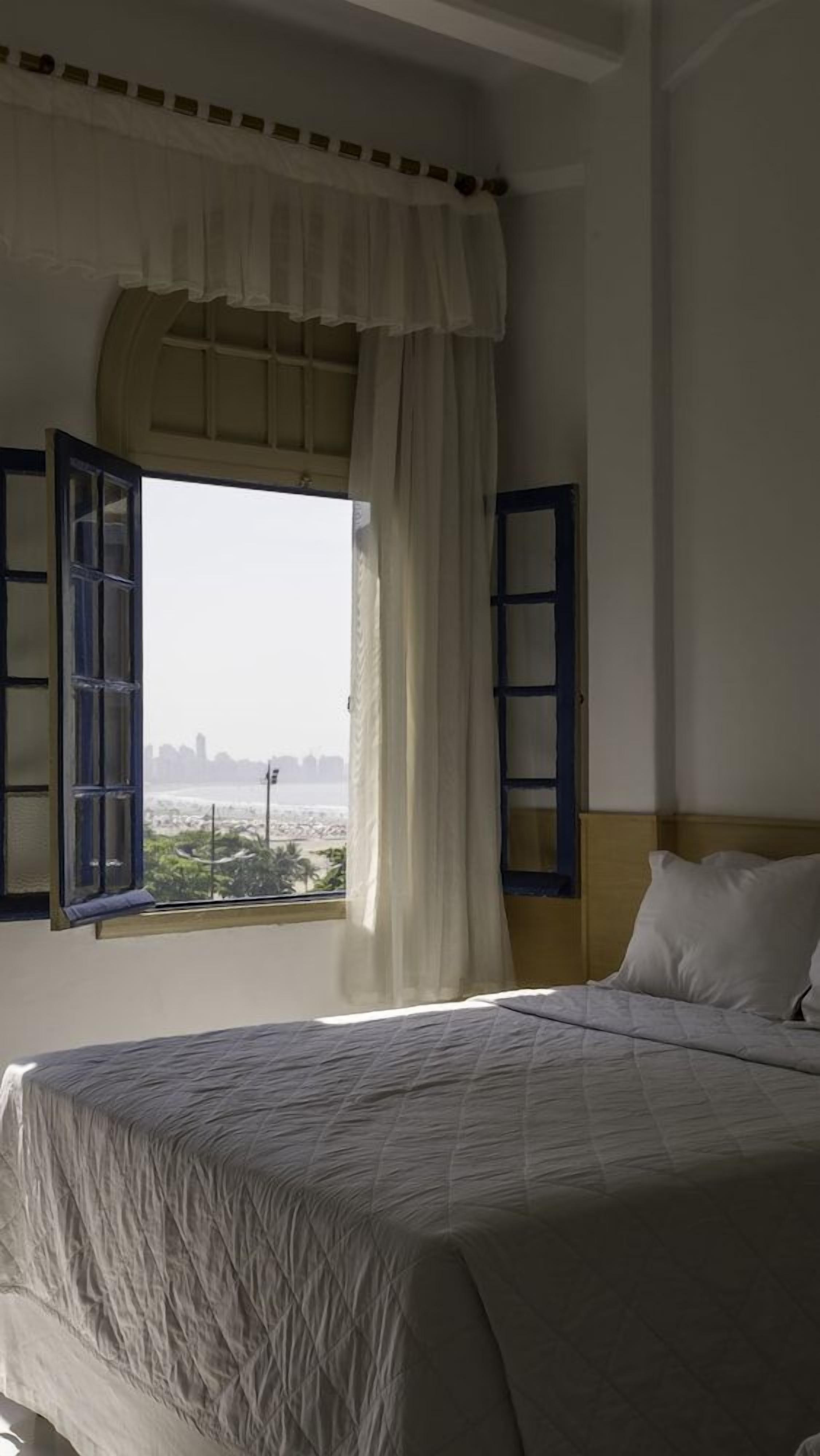 Quarto com cama, janela com cortinas e vista para a paisagem urbana.