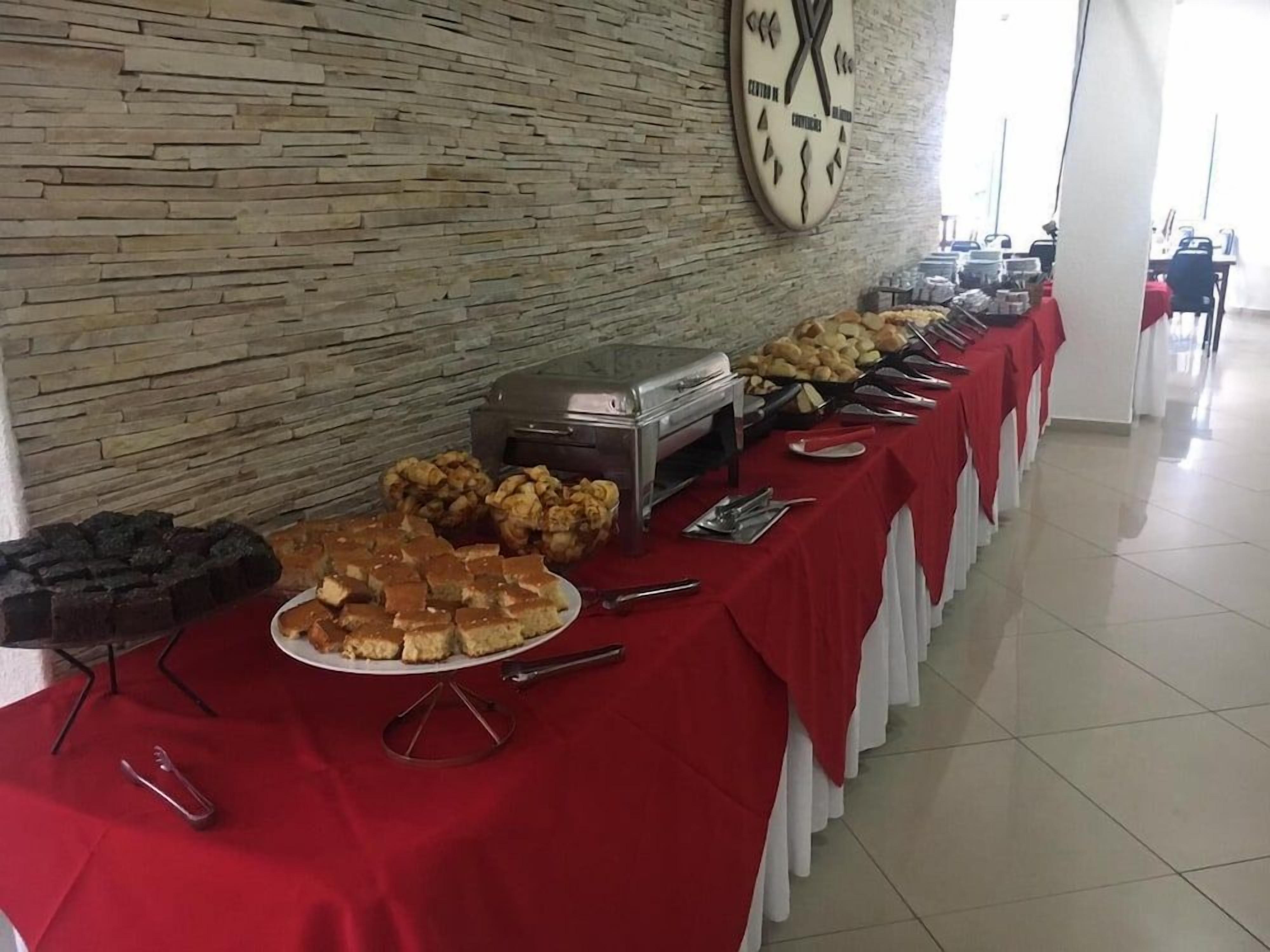 Disposição de buffet com diversos alimentos e utensílios sobre uma toalha vermelha, com uma parede de pedra e um relógio ao fundo.