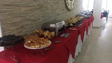 Disposição de buffet com diversos alimentos e utensílios sobre uma toalha vermelha, com uma parede de pedra e um relógio ao fundo.
