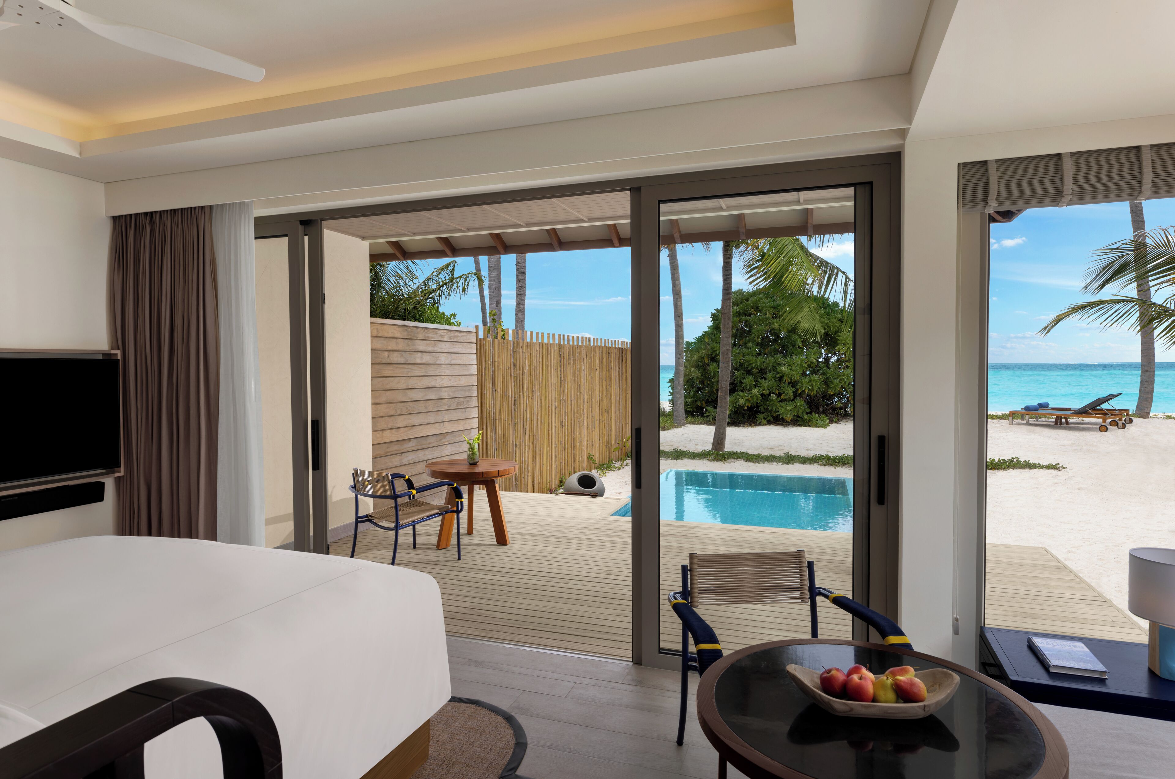 A hotel room with a bed, a TV, a dining table with fruit, and a view of a pool and beach.