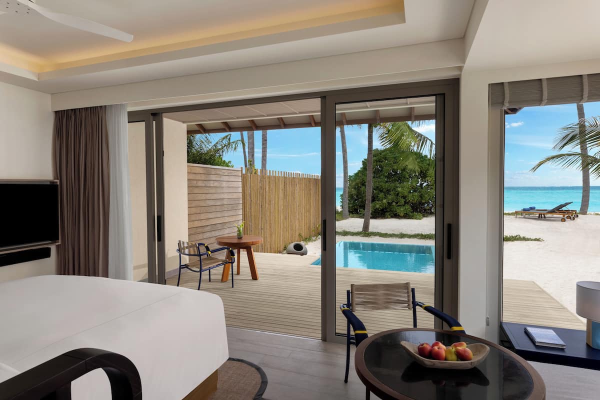 A hotel room with a bed, a TV, a dining table with fruit, and a view of a pool and beach.