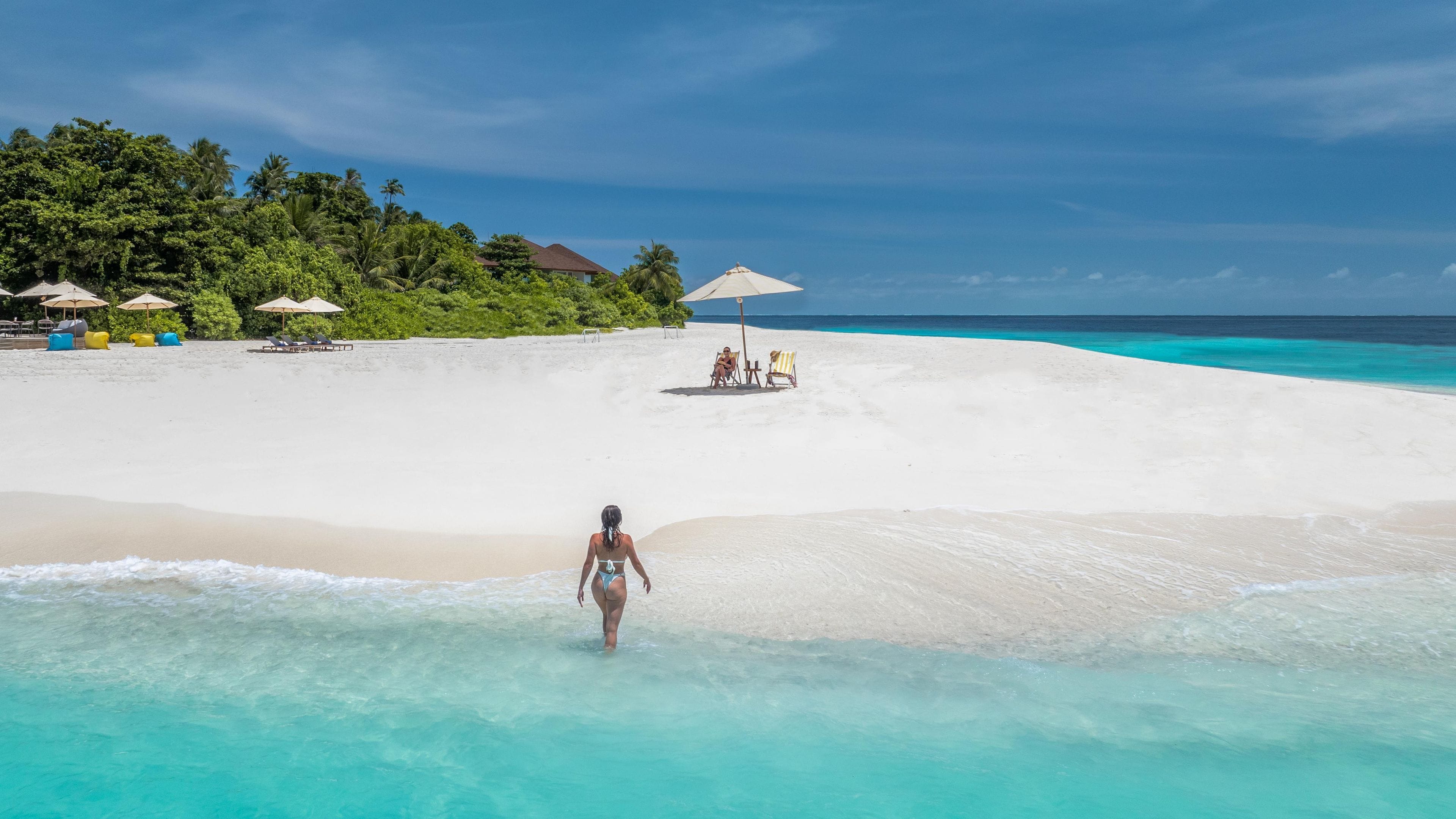 A beach with clear turquoise water, a woman walking in the water, and beach chairs and umbrellas in the background.